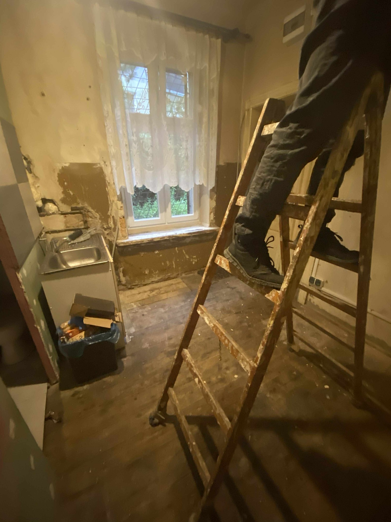 Remont starej kuchni: drabina, odsłonięte ściany, okno z firanką. Widoczny fragment zlewu i śmieci. Przygotowania do prac wykończeniowych w zaniedbanym wnętrzu.