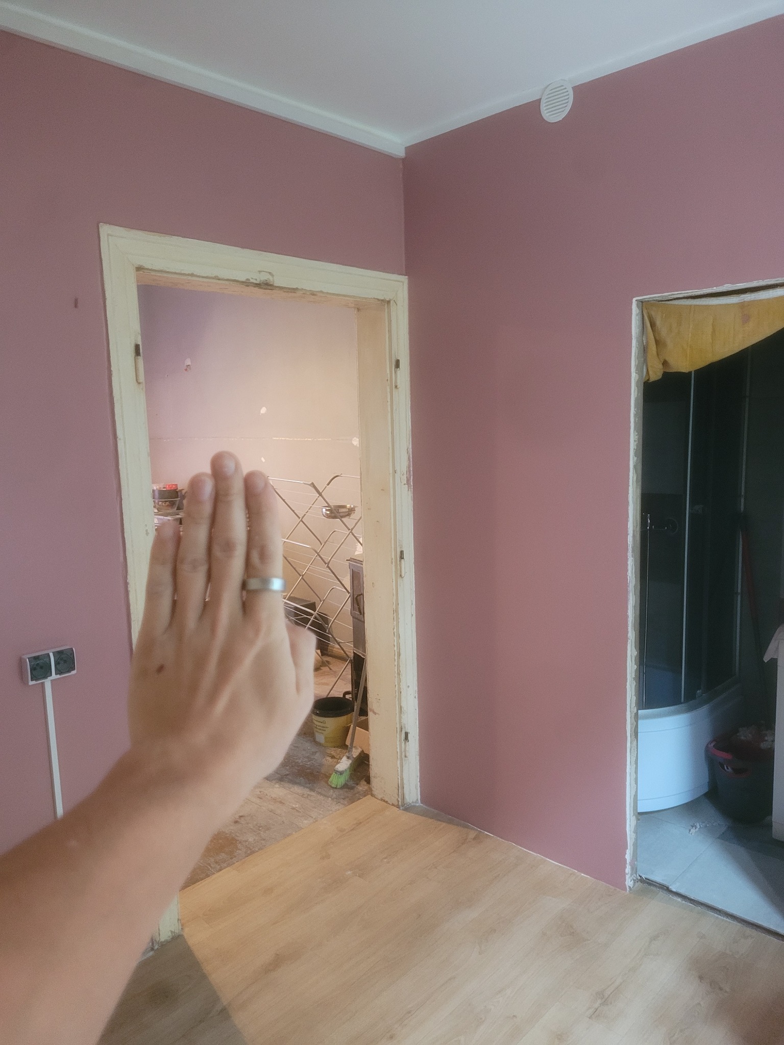 Różowa ściana z dwoma otworami drzwiowymi w trakcie wykończenia. W tle widoczna łazienka i podłoga z panelami. Ręka z pierścionkiem w kadrze.