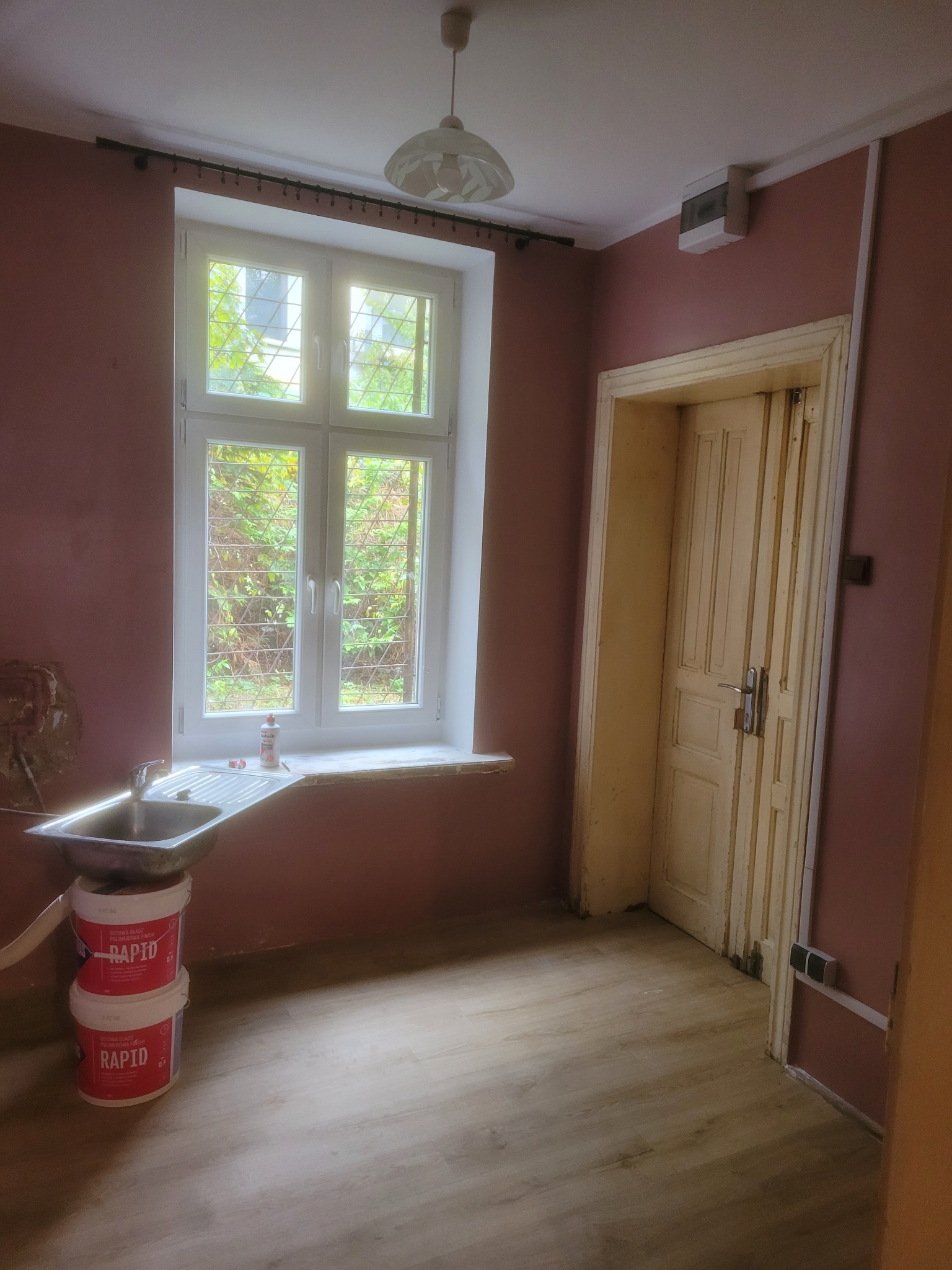 Pokój przed malowaniem: różowe ściany, okno z widokiem na zieleń, umywalka i dwa wiadra z farbą. Widoczne ślady wcześniejszych prac remontowych.