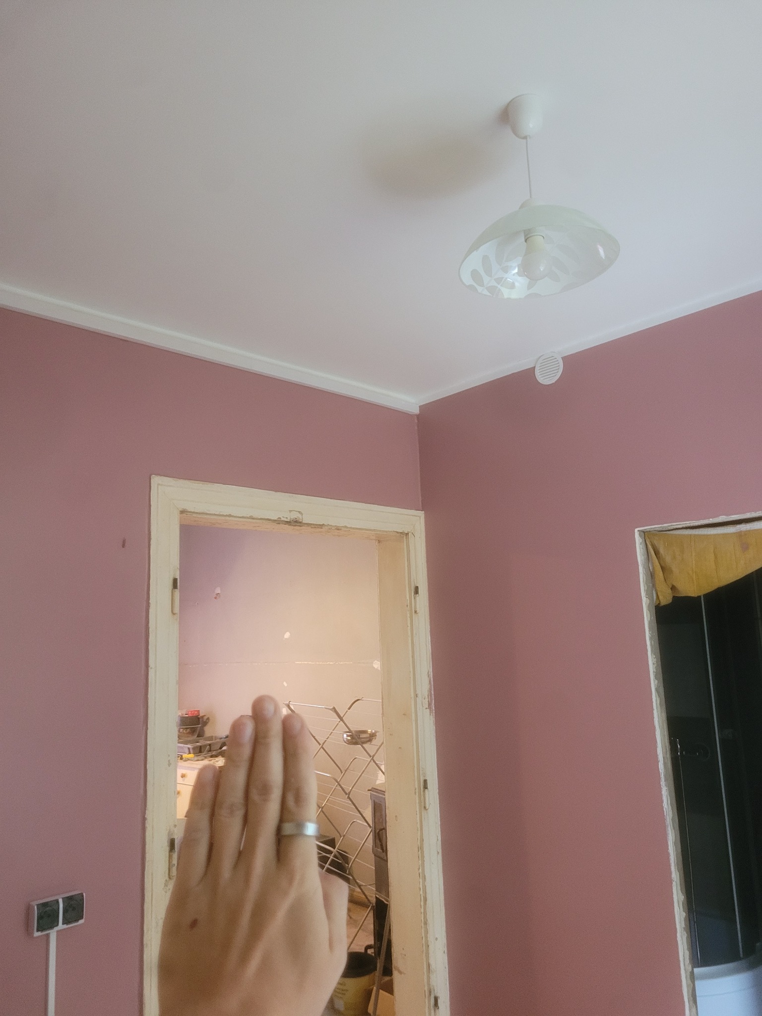 Różowa ściana po malowaniu z widocznym fragmentem dłoni w kadrze. W tle widoczne drzwi i lampa sufitowa. Całość w jasnym oświetleniu.