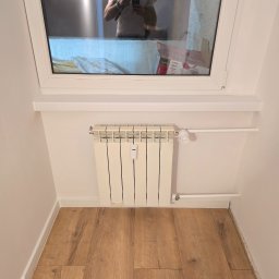 WinnerMMA - Jasne panele podłogowe w małym pokoju z grzejnikiem pod oknem, w którym odbija się osoba robiąca zdjęcie. Minimalistyczne wykończenie wnętrza.