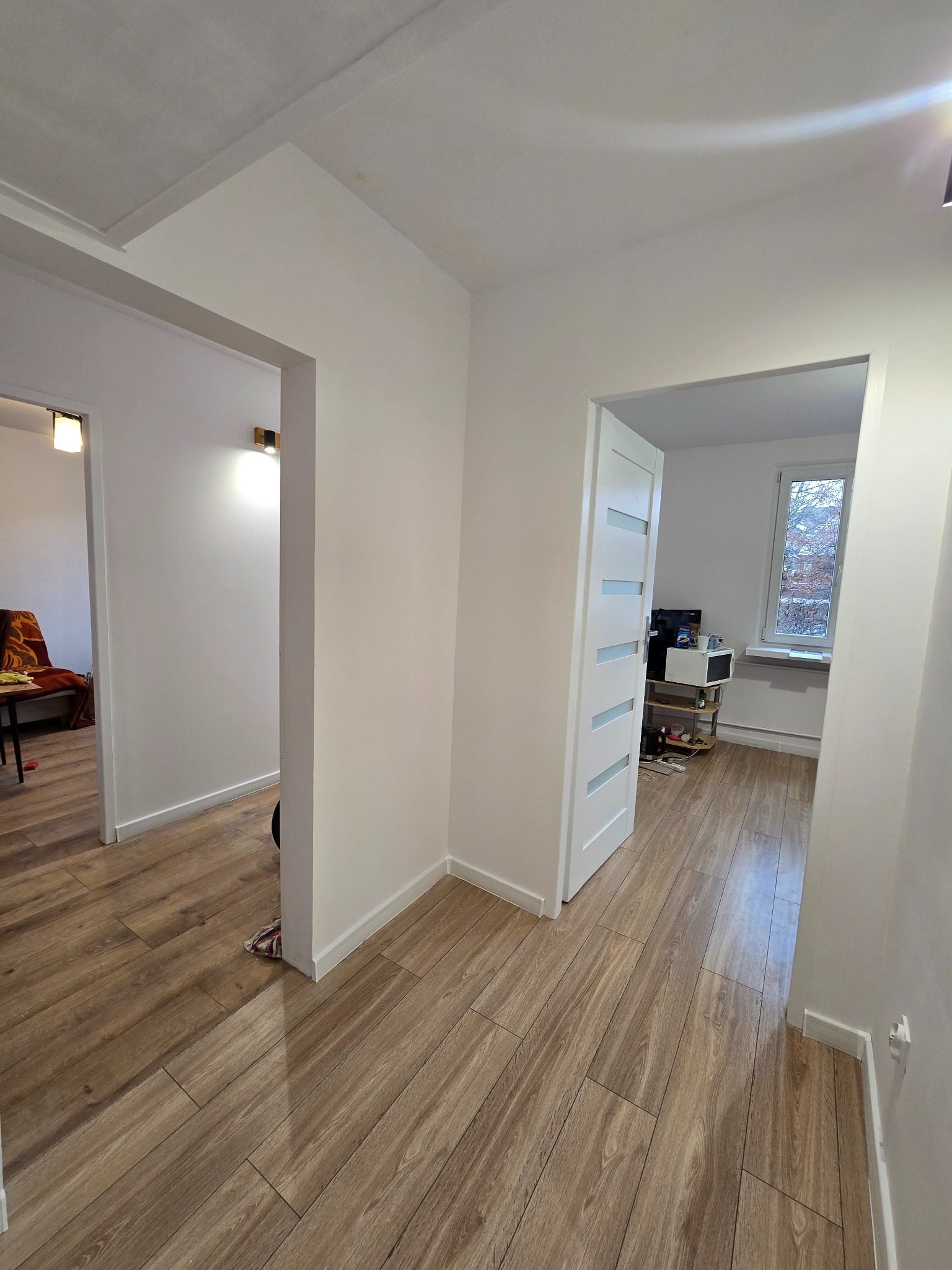 Jasne wnętrze po remoncie: drewnopodobna podłoga, białe ściany i drzwi z przeszkleniami. Widok na pokoje z oświetleniem i oknem.