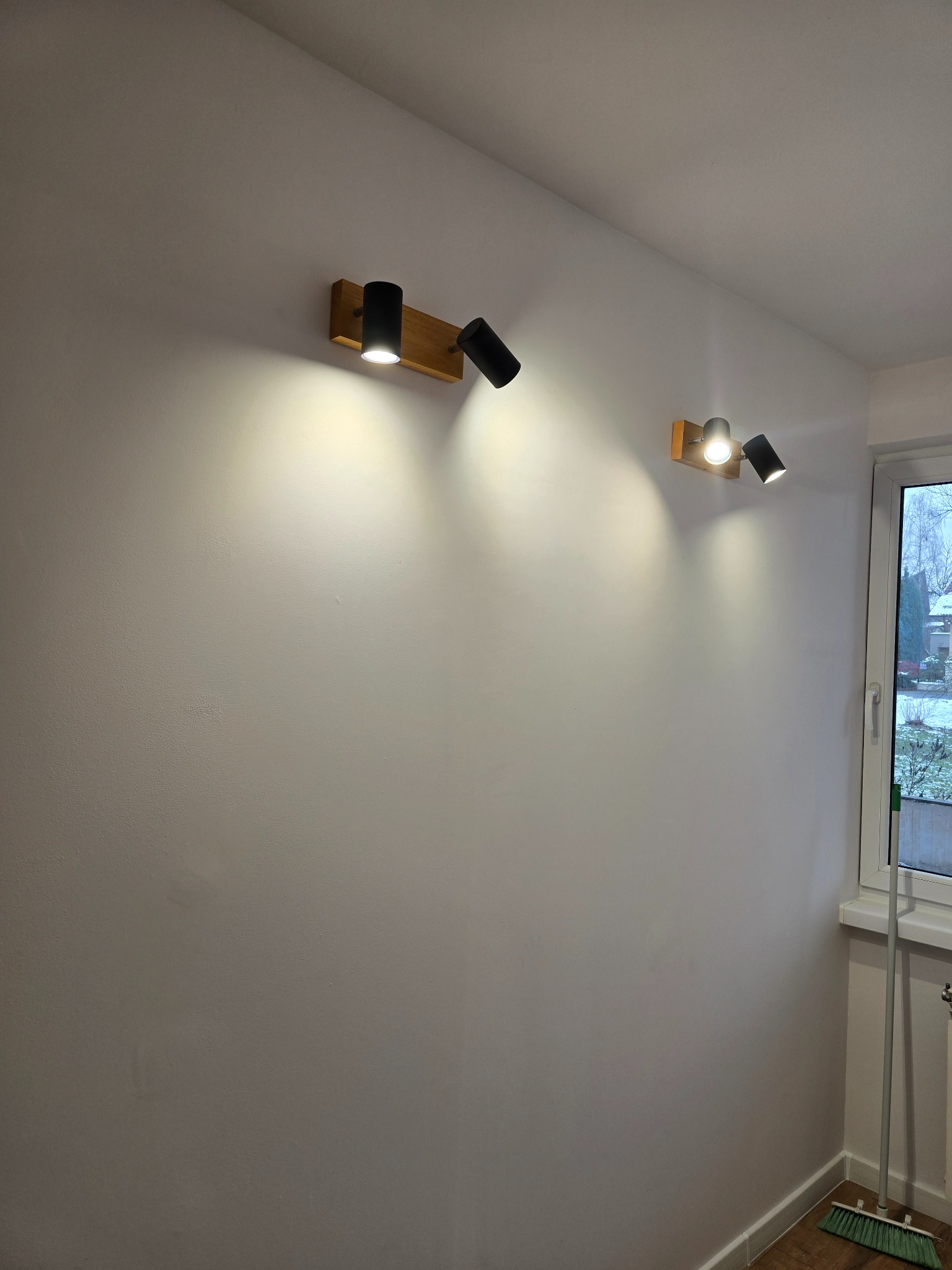 Dwa nowoczesne, regulowane reflektory LED na drewnianej listwie, zamontowane na świeżo pomalowanej, gładkiej ścianie w odcieniach szarości. Widoczne okno i fragment podłogi.
