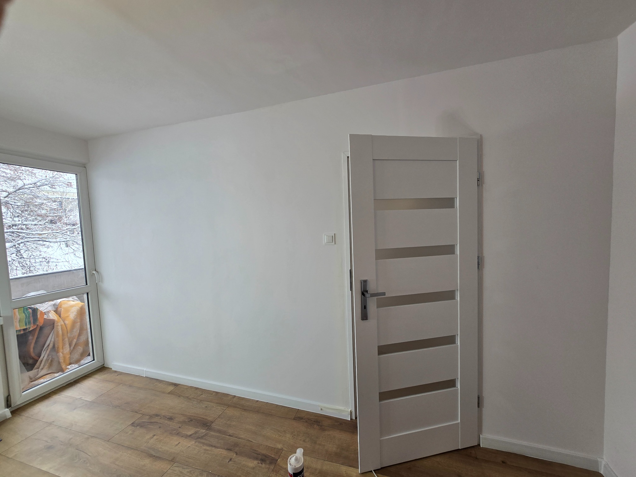 Jasne wnętrze po remoncie: drewnopodobna podłoga, białe ściany i nowoczesne, białe drzwi z przeszkleniami. Za oknem widok na zimowy krajobraz.