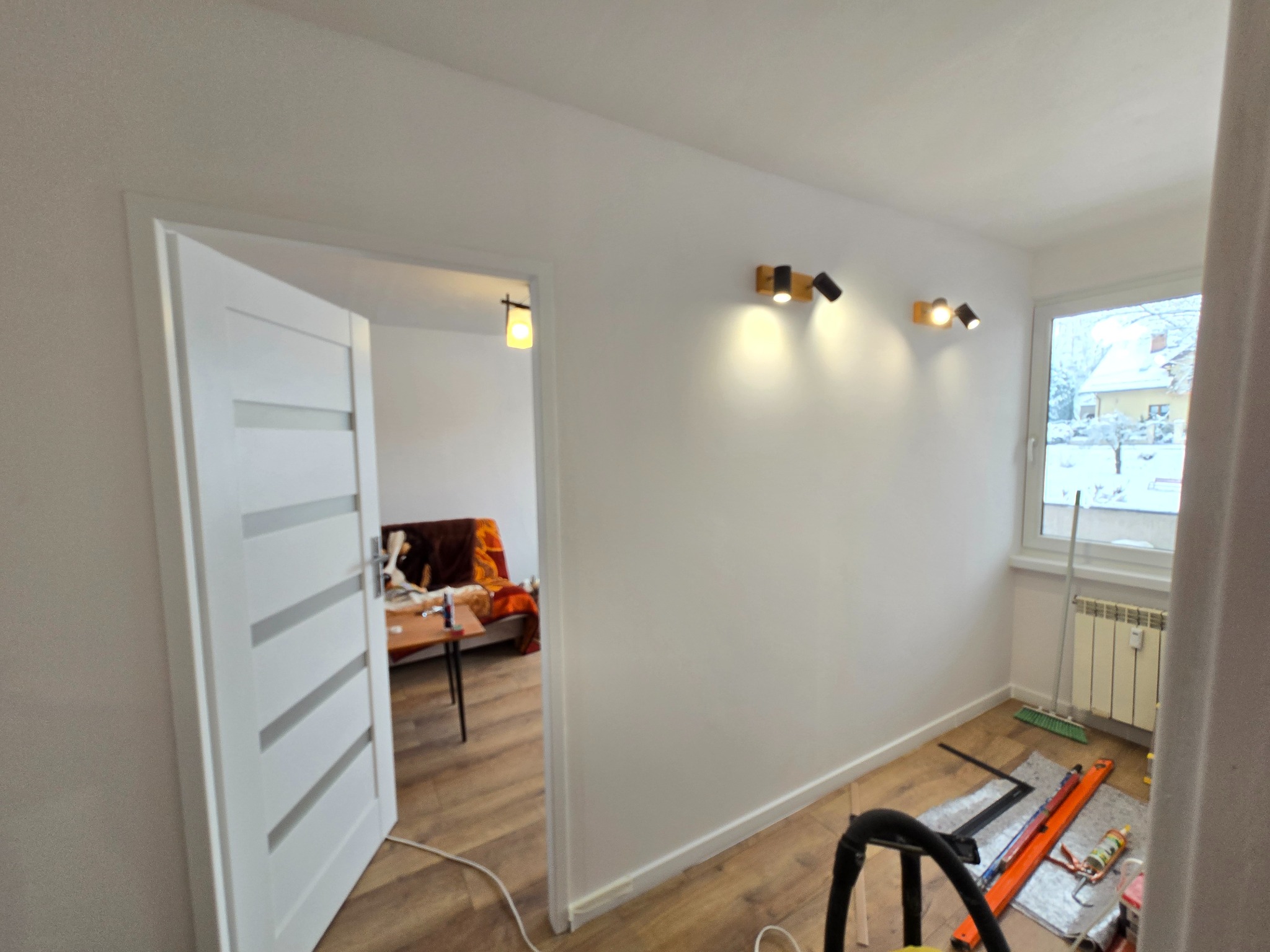 Jasne wnętrze po remoncie: białe ściany, drewniana podłoga, nowoczesne oświetlenie punktowe i białe drzwi z przeszkleniami. Widok na pokój z oknem i grzejnikiem.