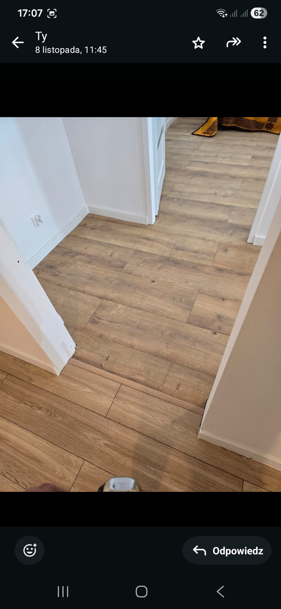 Jasne panele podłogowe ułożone w korytarzu z białymi ścianami i futrynami drzwi. Widok z góry, fragment stopy w kadrze. Nowoczesne wykończenie.