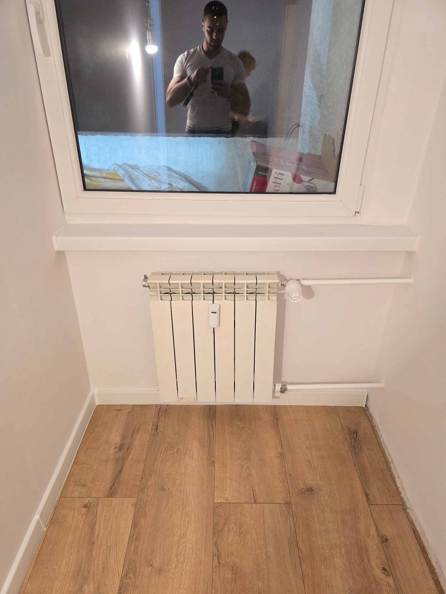 Jasne panele podłogowe w małym pokoju z grzejnikiem pod oknem, w którym odbija się osoba robiąca zdjęcie. Minimalistyczne wykończenie wnętrza.