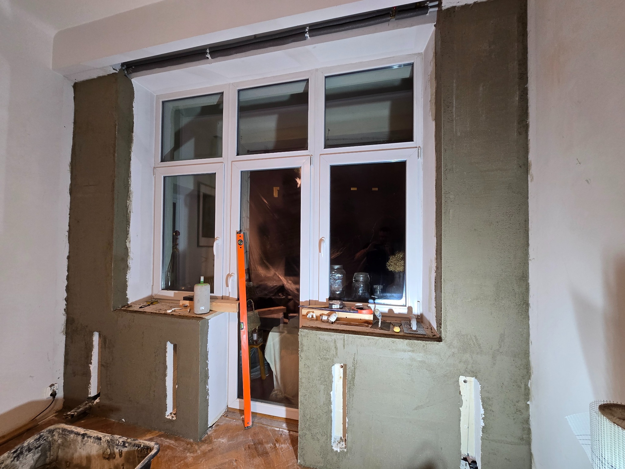 Ściana po szpachlowaniu wokół okna z widocznymi narzędziami i poziomnicą. Prace wykończeniowe w trakcie realizacji, widoczne ślady po instalacjach.