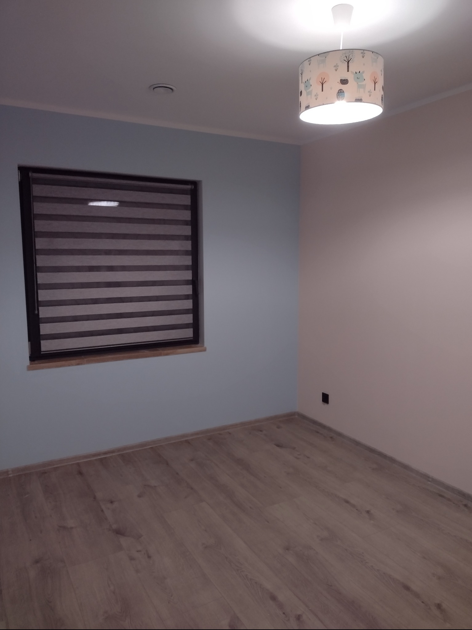 Pomalowane na dwa kolory wnętrze pokoju z drewnianą podłogą, oknem z żaluzjami i lampą sufitową z motywem zwierzęcym. Ściany w odcieniach błękitu i różu.