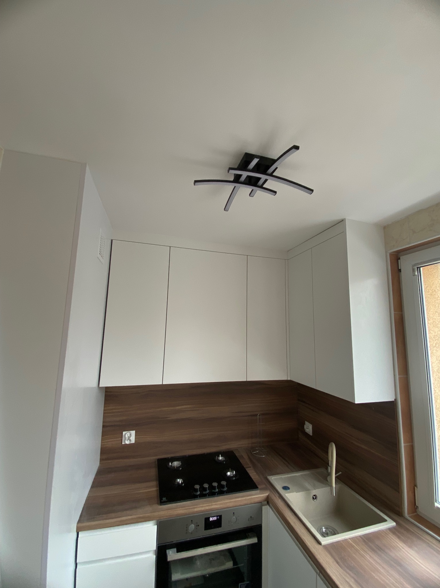 Nowoczesna kuchnia z białymi szafkami i drewnianym blatem. Czarna płyta grzewcza i designerska lampa sufitowa. Minimalistyczny styl, dbałość o detale.