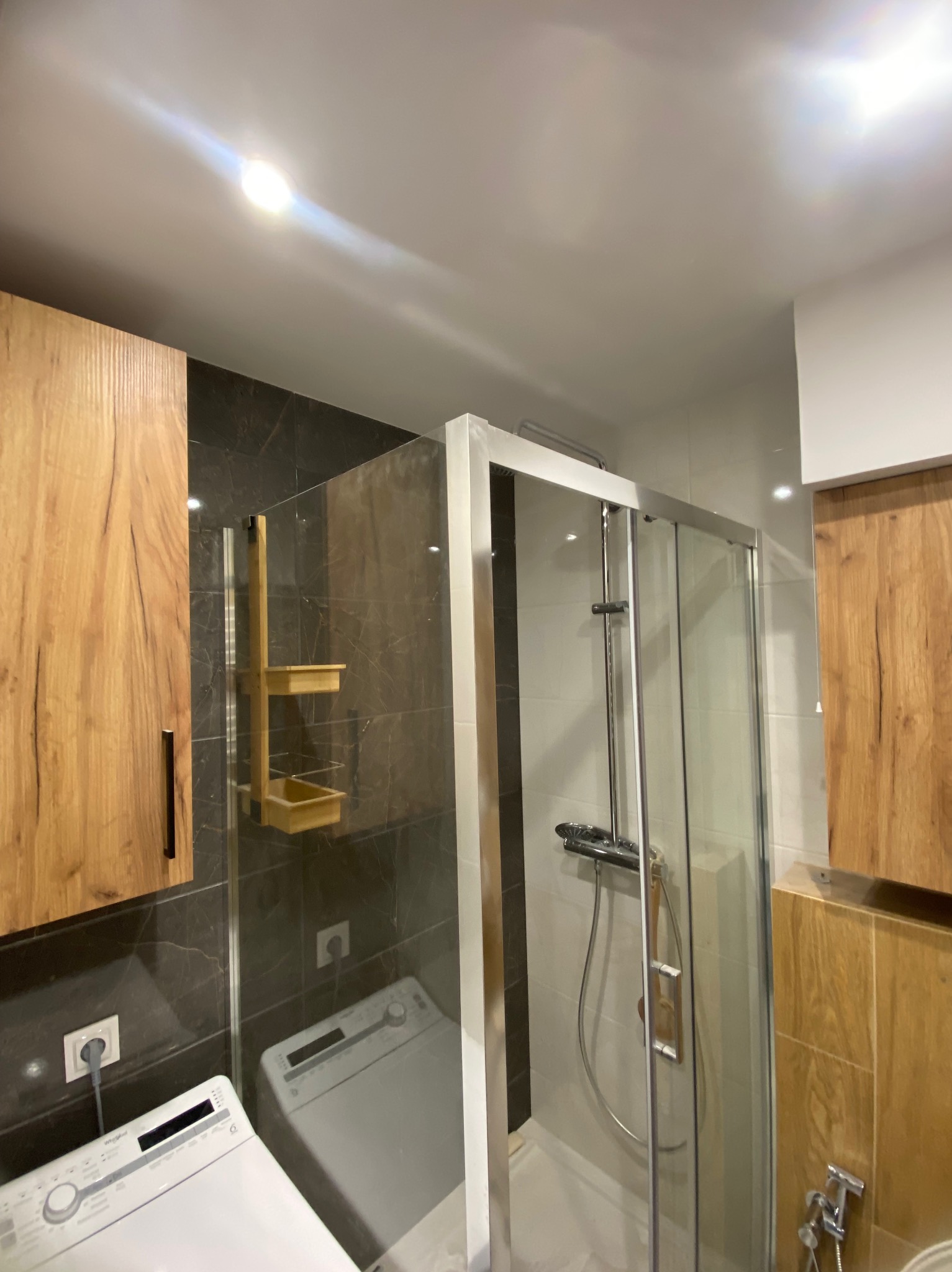Nowoczesna łazienka z kabiną prysznicową, drewnianymi szafkami i pralką. Ciemne płytki imitujące marmur na ścianie, oświetlenie sufitowe.