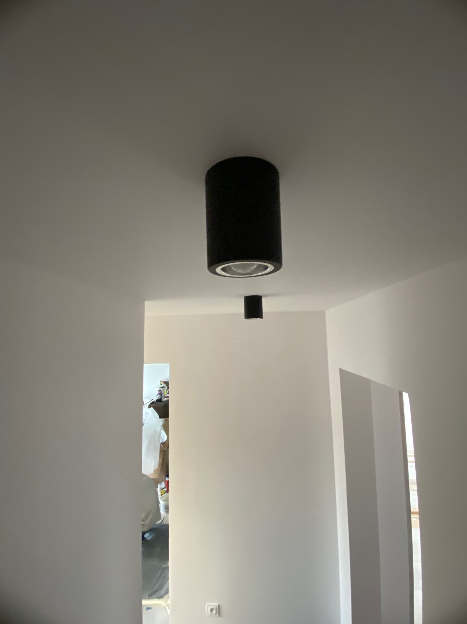 Białe wnętrze po remoncie z dwoma czarnymi lampami sufitowymi typu tuba. Widoczne świeżo pomalowane ściany i fragment sąsiedniego pomieszczenia.