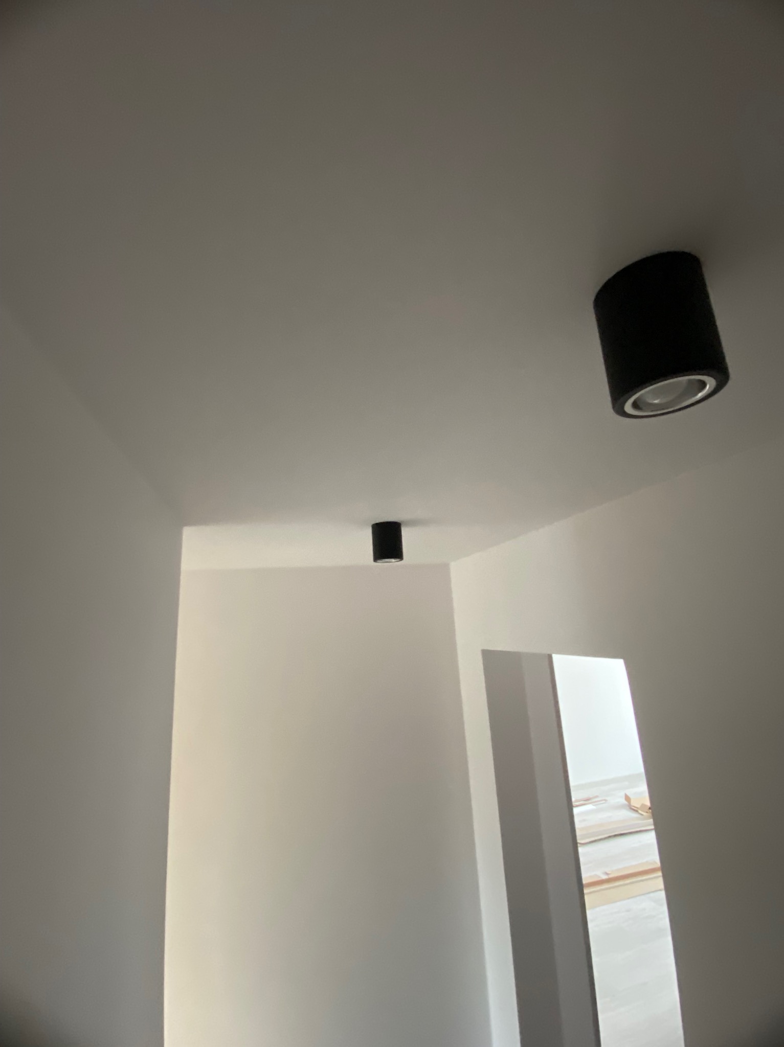 Minimalistyczne wnętrze z białymi ścianami i dwoma czarnymi, cylindrycznymi lampami sufitowymi. Widok na korytarz z materiałami budowlanymi.