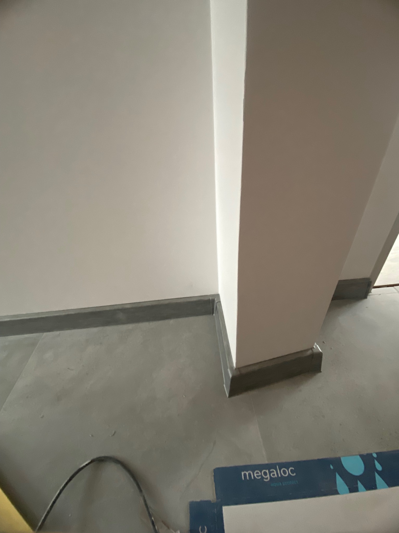 Szare płytki podłogowe i ciemne listwy przypodłogowe przy białej ścianie i filarze. Fragment opakowania paneli podłogowych Megaloc widoczny w rogu.