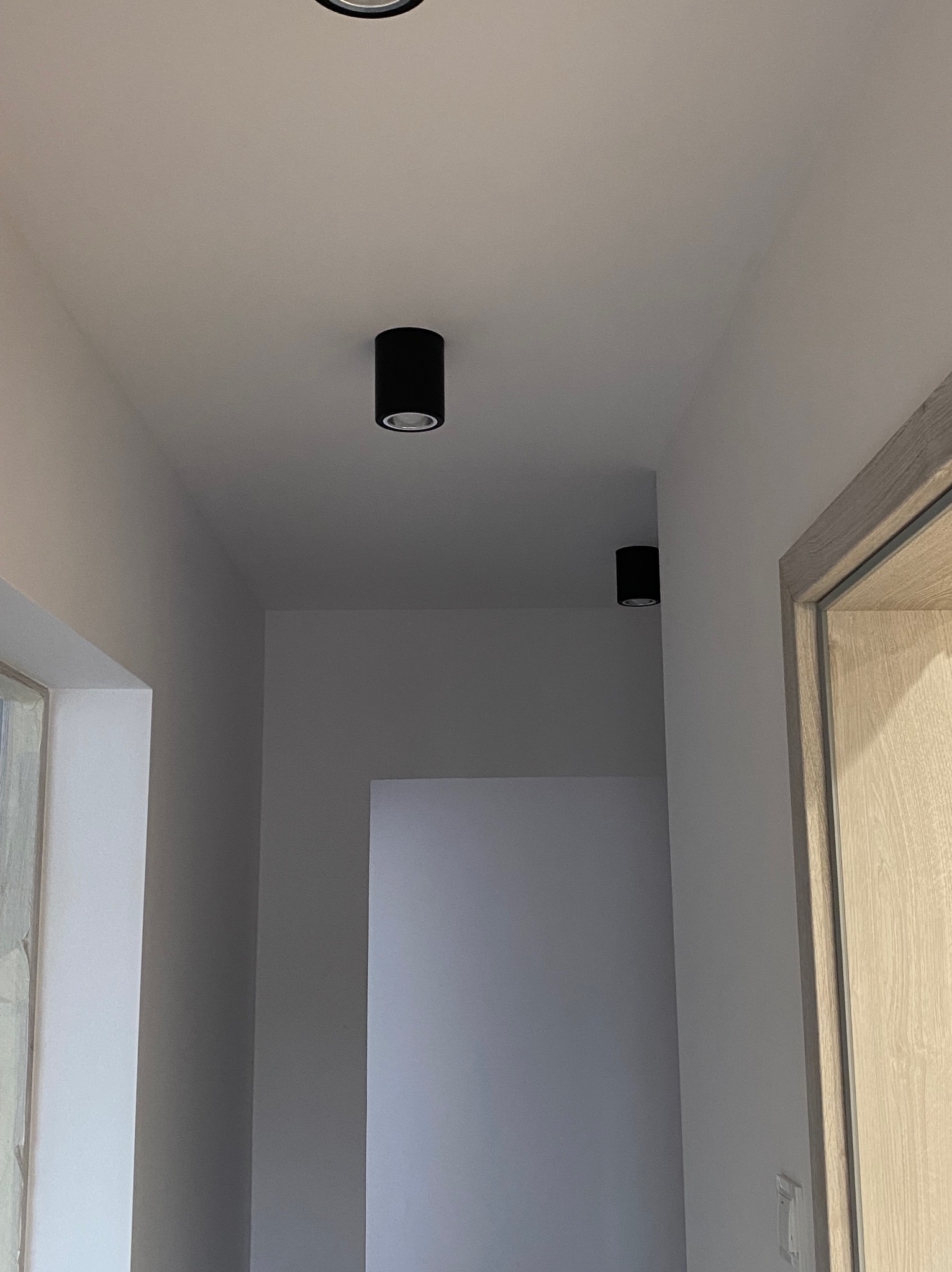 Nowoczesne, czarne lampy sufitowe typu tuba w jasnym korytarzu z widocznym fragmentem ościeżnicy drzwiowej o drewnianej fakturze.