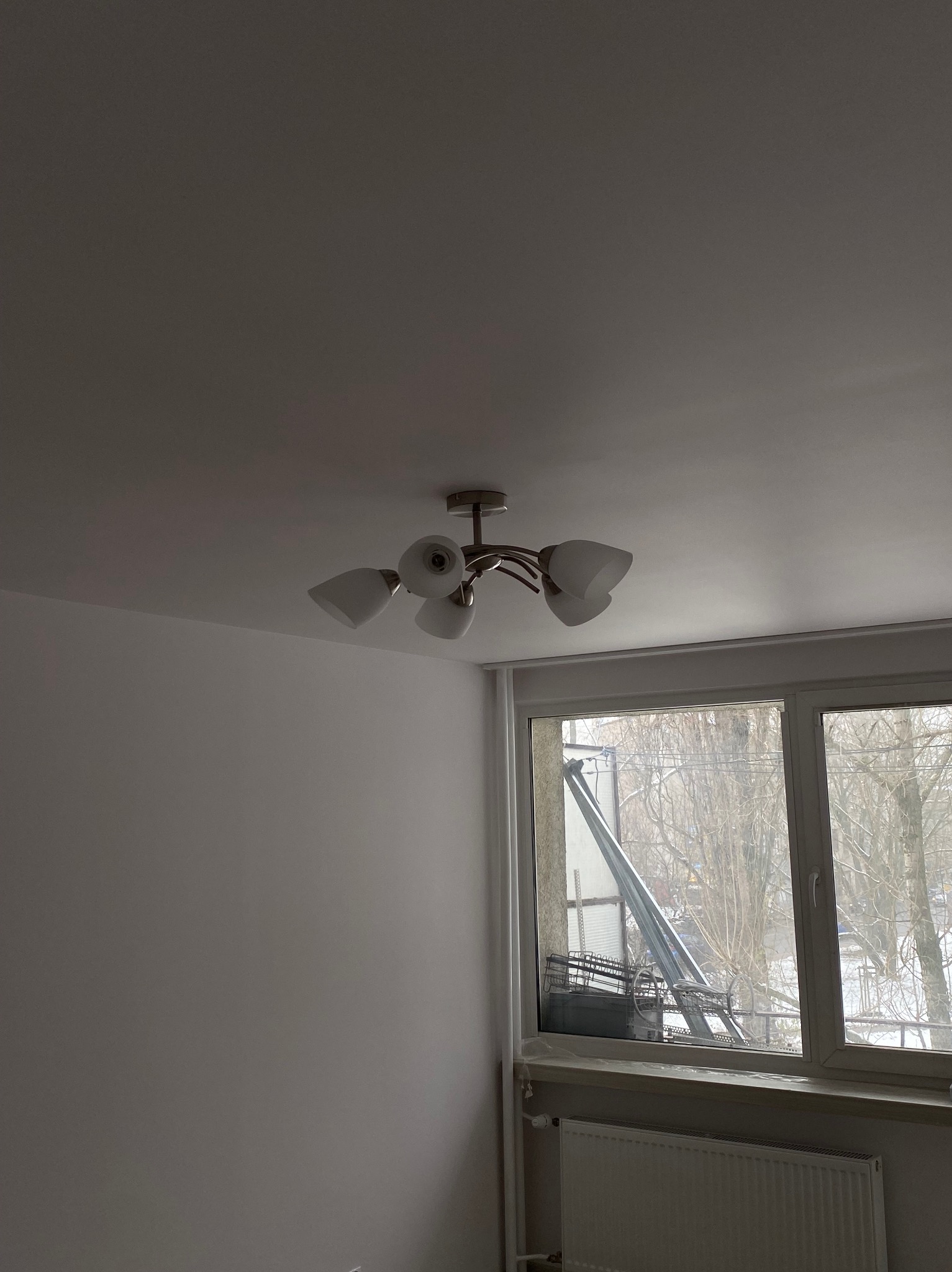 Jasne wnętrze po remoncie: świeżo pomalowane ściany, nowoczesna lampa sufitowa, okno z widokiem na drzewa. Grzejnik pod oknem. Minimalistyczny design.