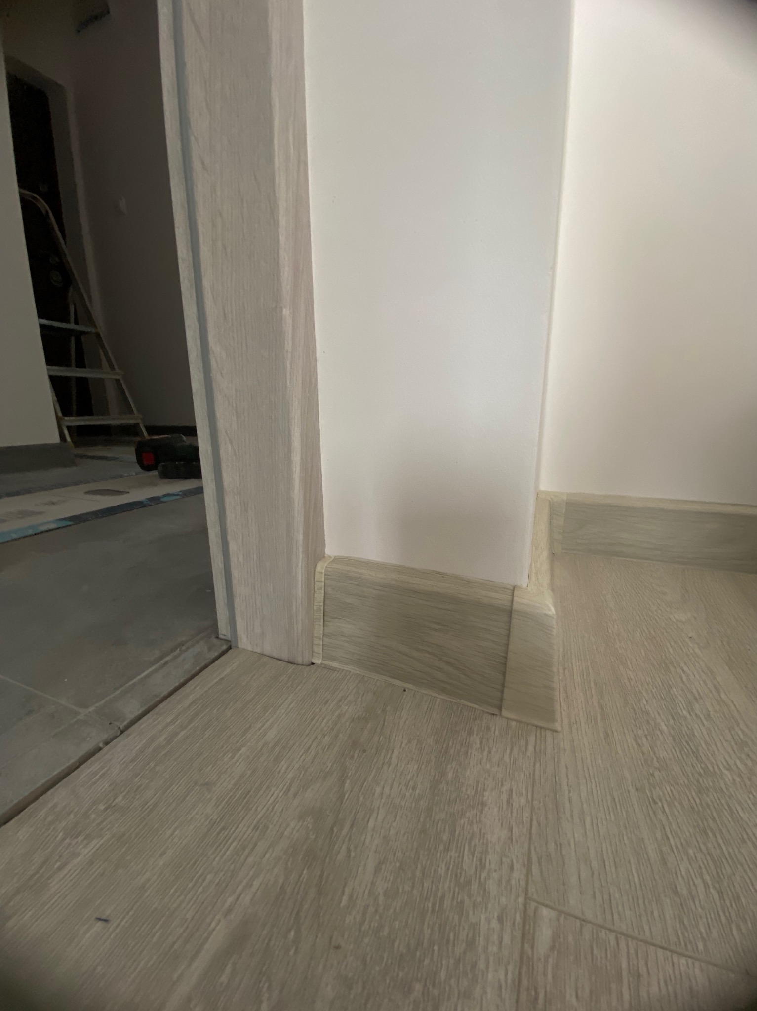 Zamknięte ujęcie na listwę przypodłogową i ościeżnicę drzwiową w odcieniach szarości, z widocznym fragmentem podłogi i ściany. W tle drabina.