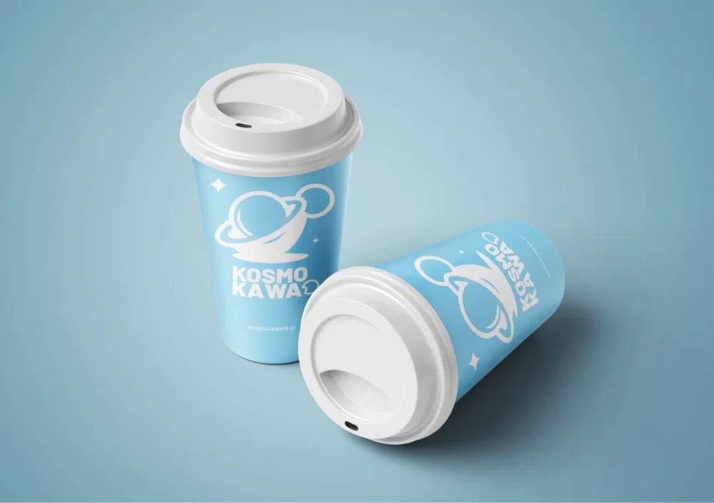 Dwa błękitne kubki z białymi pokrywkami, z logo 'Kosmo Kawa' z motywem planety z uszkami, leżący i stojący, na jednolitym, błękitnym tle.