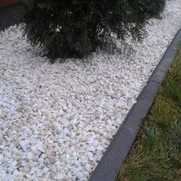 JIRASOFT GROUP SPÓŁKA Z OGRANICZONĄ ODPOWIEDZIALNOŚCIĄ - Białe kamienie dekoracyjne w ogrodzie, obok trawnika i małego drzewka iglastego. Granica oddzielona betonowym obrzeżem. Widok z góry, bliski kadr.