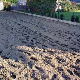 JIRASOFT GROUP SPÓŁKA Z OGRANICZONĄ ODPOWIEDZIALNOŚCIĄ - Przygotowany teren pod trawnik z widocznymi śladami stóp, ogrodzeniem i młodymi tujami w tle. Słoneczny dzień, widok z perspektywy ziemi.