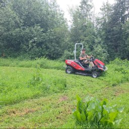 JIRASOFT GROUP SPÓŁKA Z OGRANICZONĄ ODPOWIEDZIALNOŚCIĄ - Czerwona kosiarka samojezdna marki SECO w trakcie koszenia wysokiej trawy na zarośniętej działce. Widoczny operator i częściowo skoszony teren.