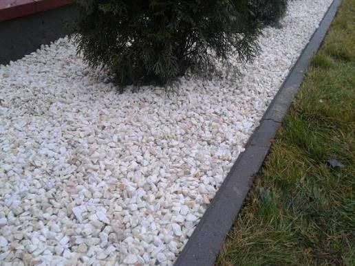 Białe kamienie dekoracyjne w ogrodzie, obok trawnika i małego drzewka iglastego. Granica oddzielona betonowym obrzeżem. Widok z góry, bliski kadr.