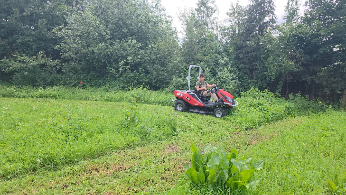 Czerwona kosiarka samojezdna marki SECO w trakcie koszenia wysokiej trawy na zarośniętej działce. Widoczny operator i częściowo skoszony teren.
