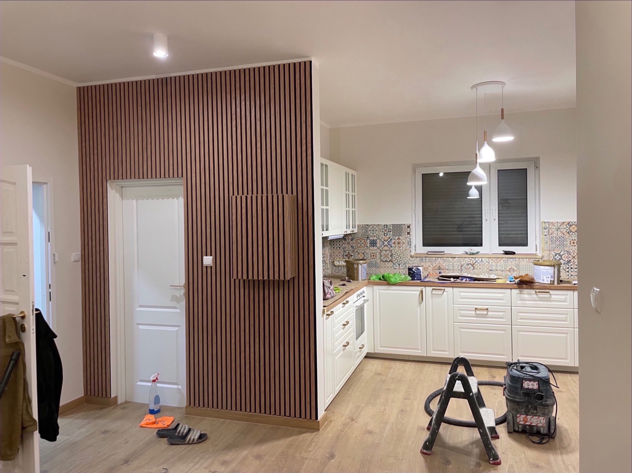 Nowoczesna aranżacja wnętrza: drewniane lamele na ścianie, biała kuchnia z wzorzystą mozaiką nad blatem, drabina i odkurzacz na podłodze.