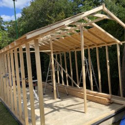 Marek - Konstrukcja szkieletowa budynku w trakcie budowy, widoczne drewniane elementy, drabiny i narzędzia na betonowej podstawie, otoczone zielenią.