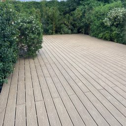 Marek - Jasna, drewniana podłoga tarasu z kompozytu, otoczona zielenią krzewów i drzew. Deski ułożone równolegle, tworzą uporządkowaną przestrzeń. Widok na zewnątrz, w tle roślinność.