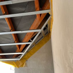 Marek - Montaż metalowego stelażu sufitu podwieszanego, widoczne drewniane elementy konstrukcyjne i folia ochronna przy ścianie. Ujęcie z dołu, sufit w trakcie prac.