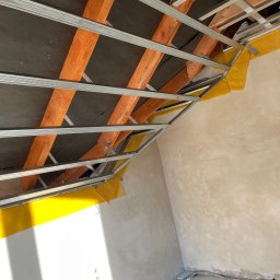 Marek - Montaż metalowego stelażu sufitu podwieszanego z płyt karton-gips, widoczne drewniane elementy konstrukcyjne i żółta folia izolacyjna. Ściany w trakcie szpachlowania.