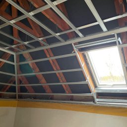 Marek - Szkielet sufitu podwieszanego z profili metalowych, widok z dołu, okno dachowe w tle. Konstrukcja na poddaszu z drewnianymi elementami.