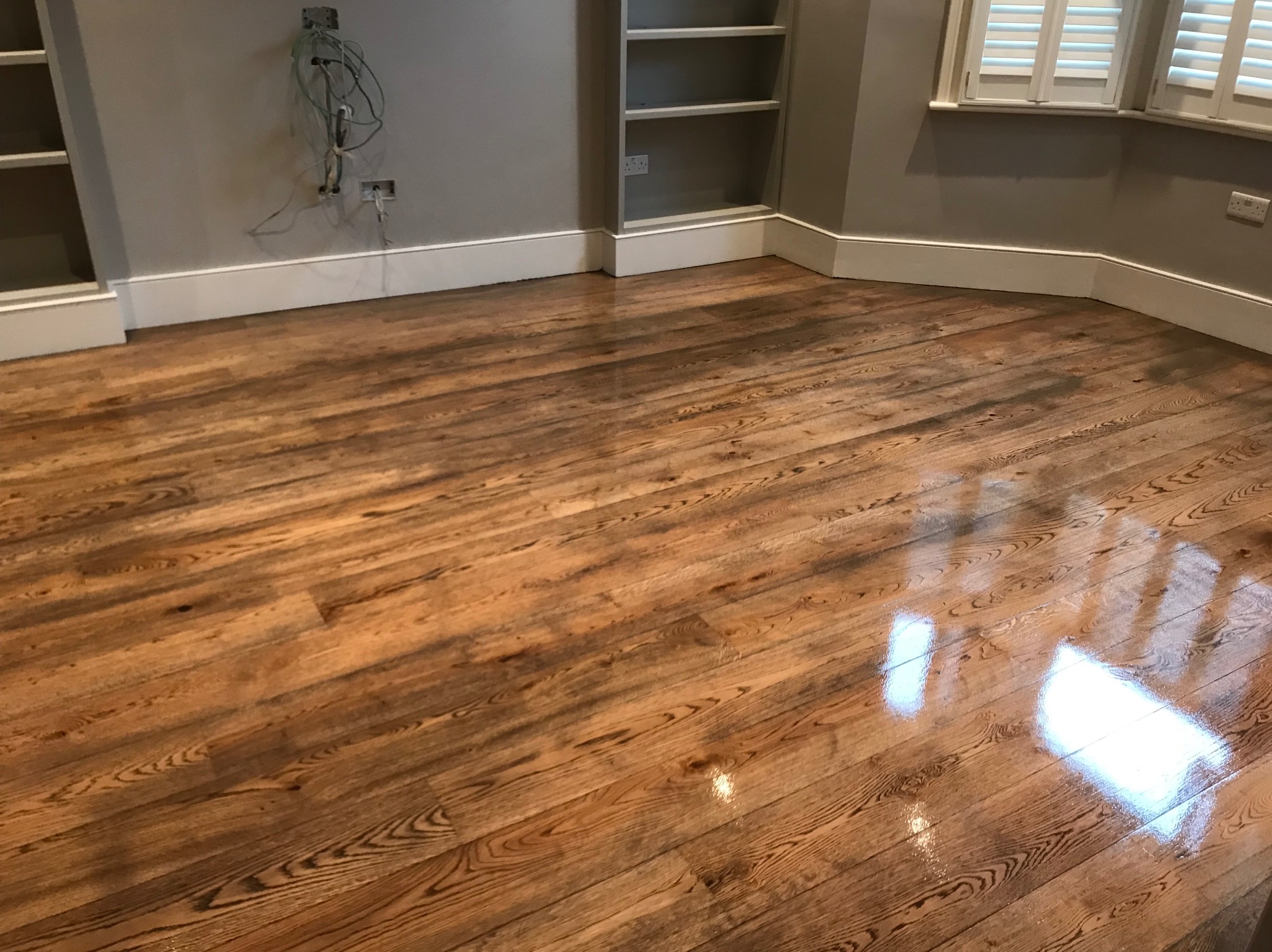 Błyszcząca, świeżo położona drewniana podłoga w pokoju z szarymi ścianami i białymi listwami przypodłogowymi. Widoczne półki wnękowe i okno z żaluzjami.