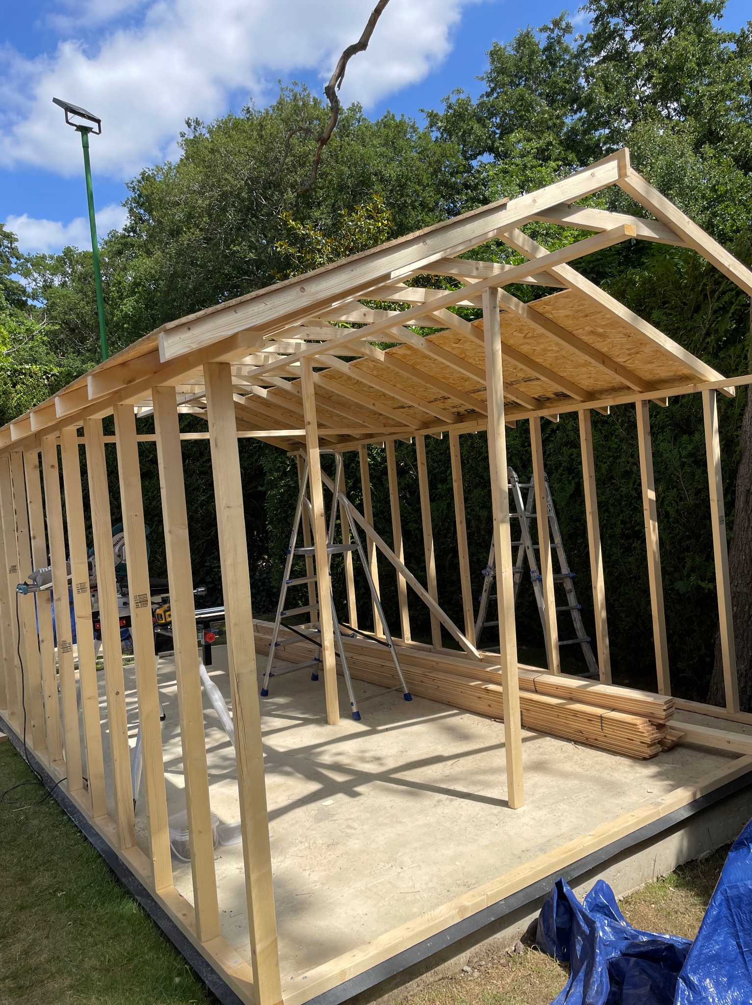 Konstrukcja szkieletowa budynku w trakcie budowy, widoczne drewniane elementy, drabiny i narzędzia na betonowej podstawie, otoczone zielenią.