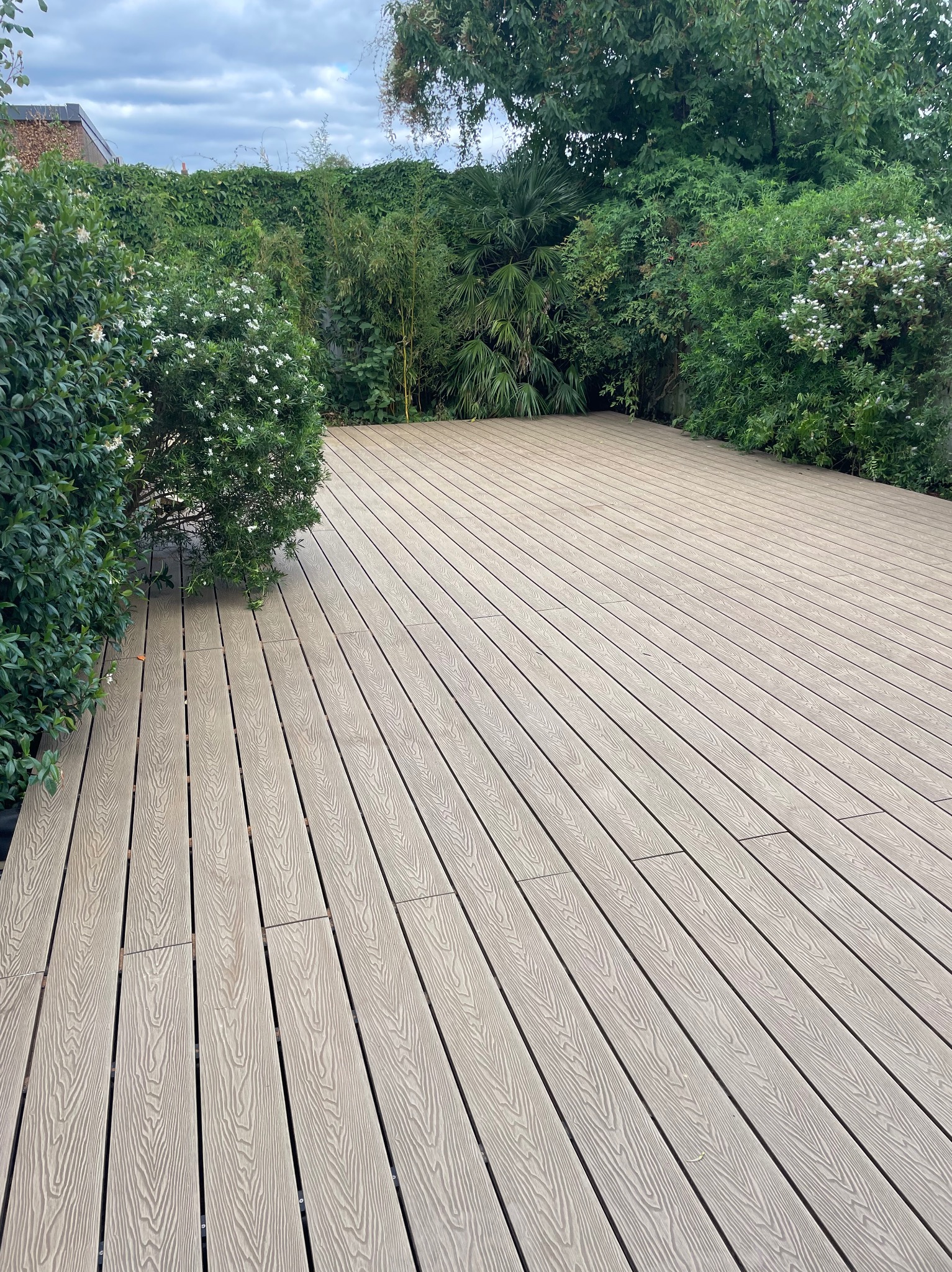 Jasna, drewniana podłoga tarasu z kompozytu, otoczona zielenią krzewów i drzew. Deski ułożone równolegle, tworzą uporządkowaną przestrzeń. Widok na zewnątrz, w tle roślinność.