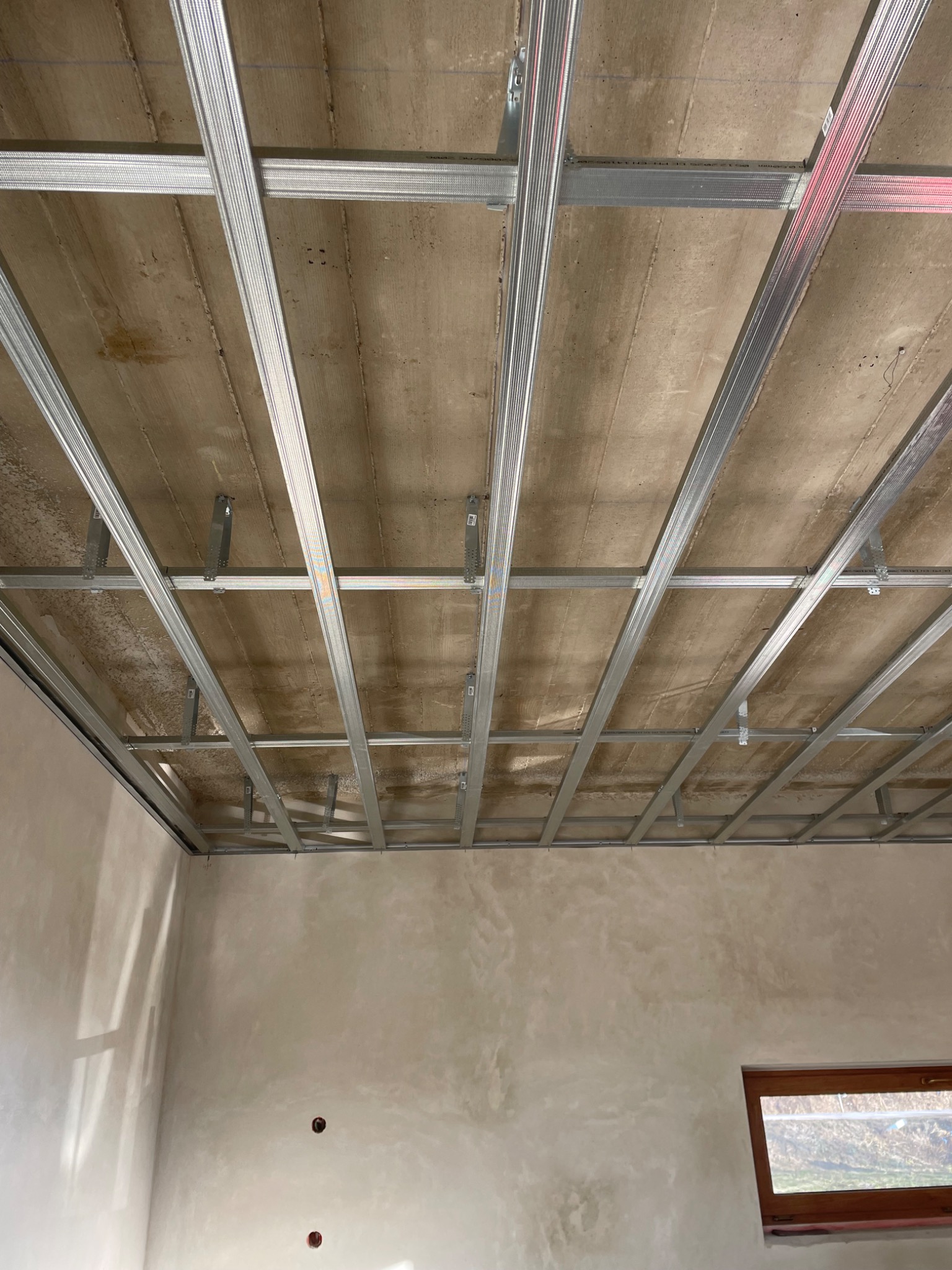 Montaż metalowej konstrukcji sufitu podwieszanego w surowym wnętrzu. Widoczne profile i płyty. Ściany w jasnym odcieniu, okno w tle.