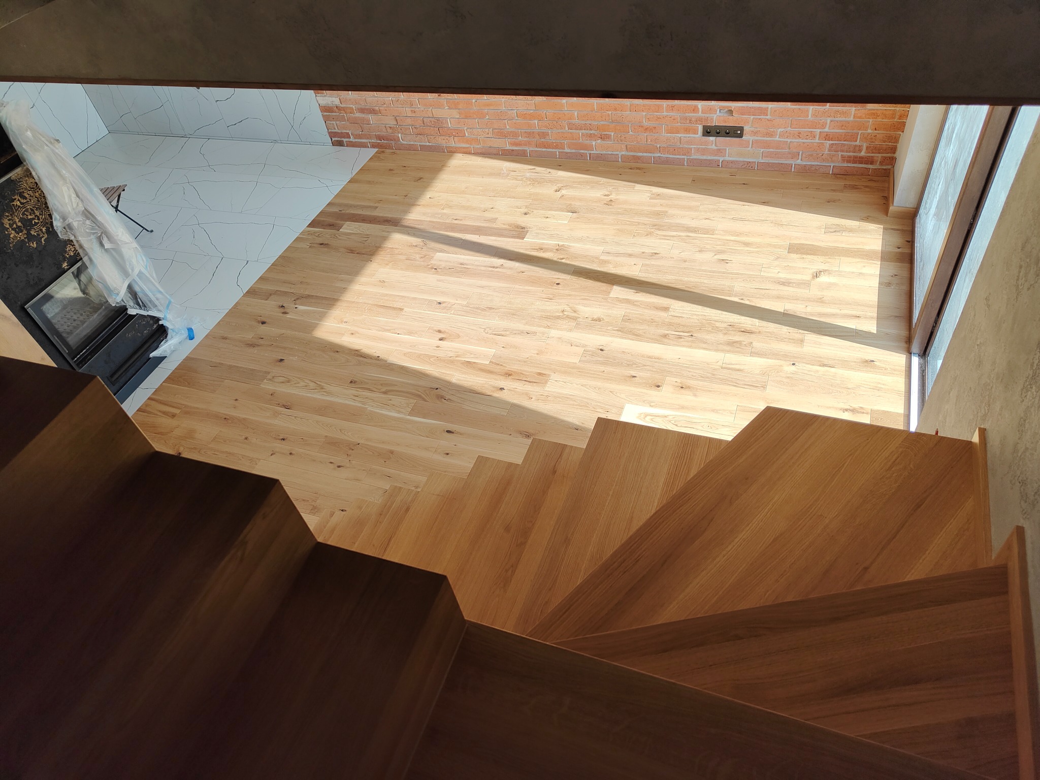 Jasny parkiet ułożony w salonie z ceglaną ścianą, widok z góry ze schodów. Część podłogi wyłożona białymi płytkami. Minimalistyczny design.