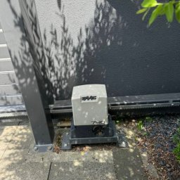 BARTŁOMIEJ PAWLIKOWSKI BER-NET - Zamontowany napęd FAAC 740 do bramy przesuwnej, widoczna listwa zębata, szara ściana z cieniem drzewa w tle. Element automatyki bram w zbliżeniu.