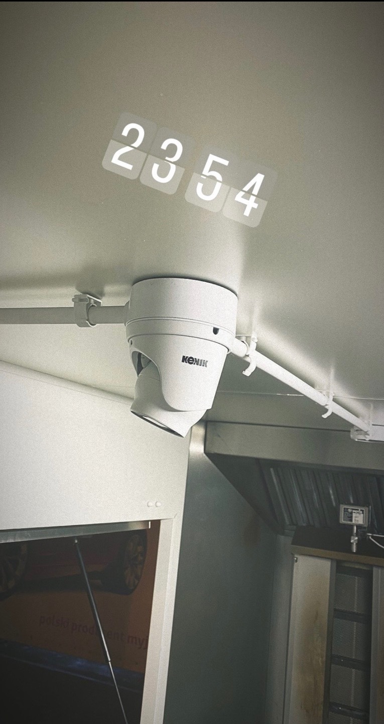 Biała kamera przemysłowa marki Konik zamontowana pod sufitem, z widocznym wyświetlaczem czasu '23:54'. Widoczne elementy wyposażenia lokalu.