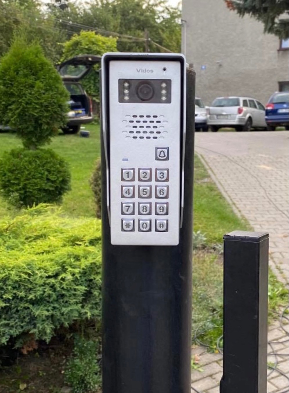 Zewnętrzny panel wideodomofonu Vidos z kamerą i klawiaturą numeryczną, zamontowany na czarnym słupku, na tle zieleni i chodnika.