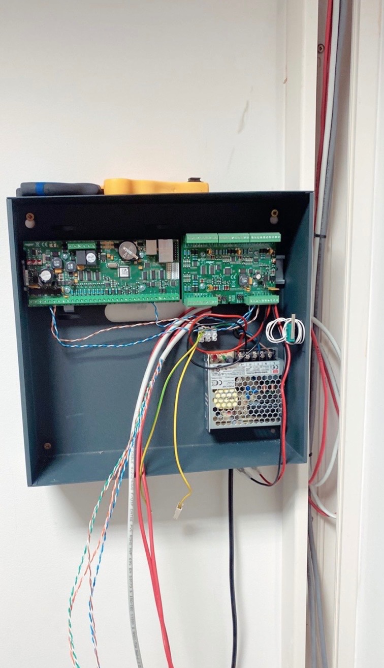 Szara skrzynka z elektroniką i kablami na białej ścianie. Widoczne płytki drukowane, zasilacz i kolorowe przewody. Instalacja alarmowa w trakcie montażu.