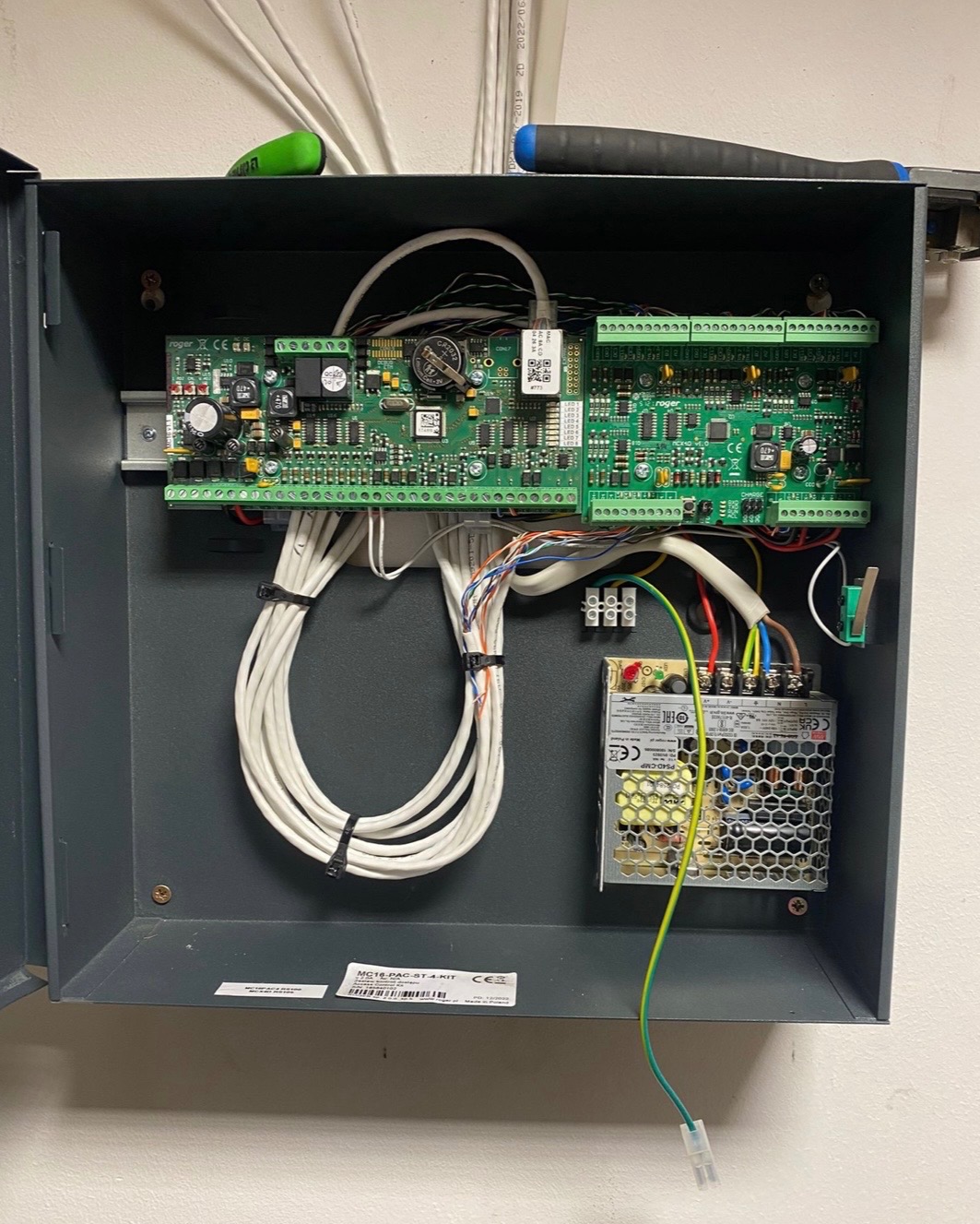 Otwarta szafka sterownicza z elektroniką alarmową: płytki drukowane, zasilacz impulsowy, okablowanie. Widoczne elementy montażowe i oznaczenia producenta.