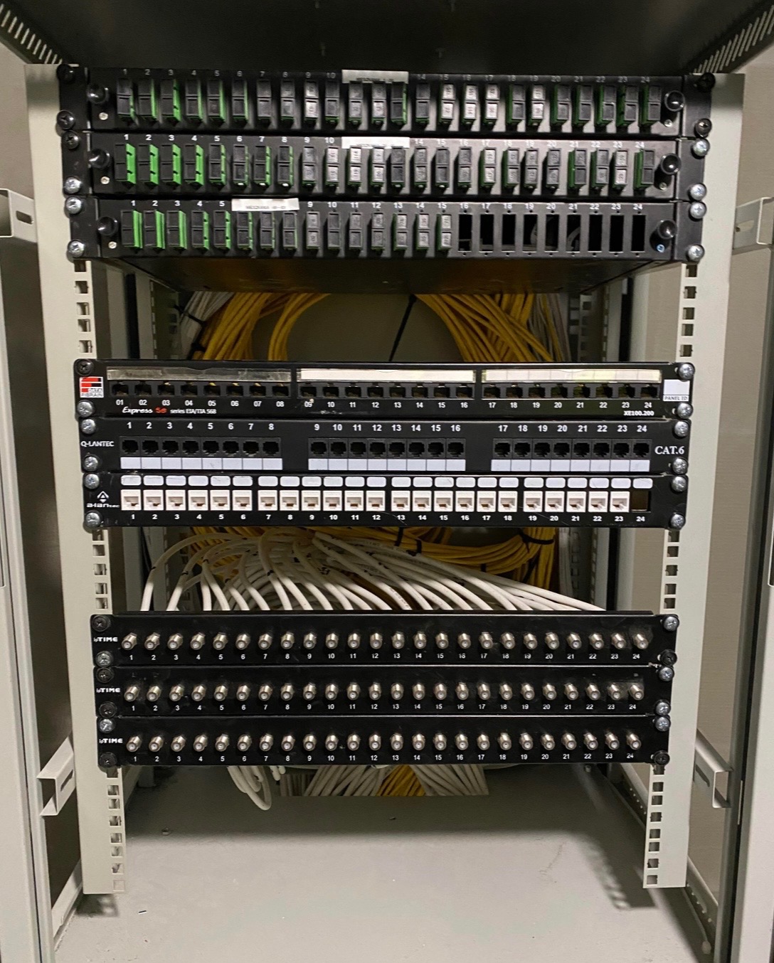 Panel krosowniczy CAT.6 w szafie rack, z widocznymi kablami Ethernet w kolorach białym i żółtym. Złącza ponumerowane, etykiety na panelach. Widok z bliska na okablowanie.