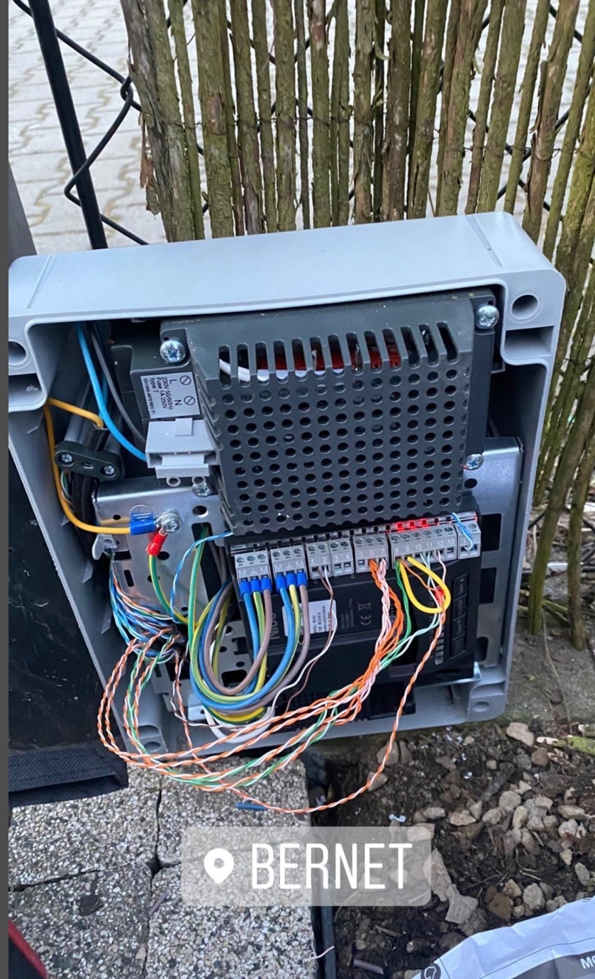 Otwarta szara skrzynka sterownicza z podłączonymi przewodami, widoczna na zewnątrz, na tle ogrodzenia z gałęzi. Instalacja elektryczna w trakcie montażu.