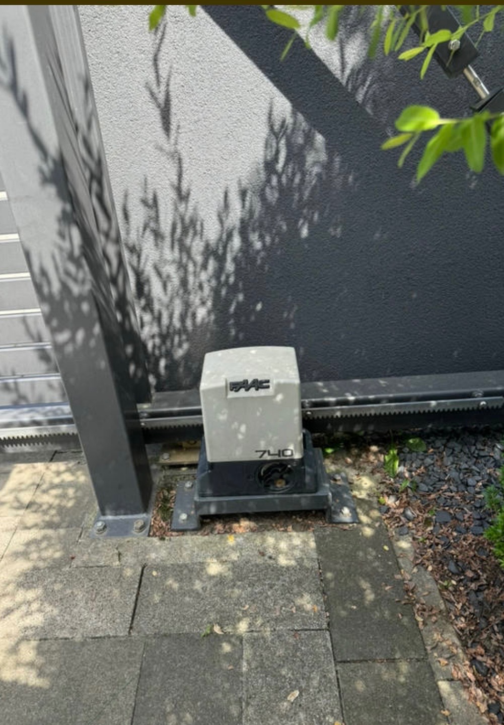 Zamontowany napęd FAAC 740 do bramy przesuwnej, widoczna listwa zębata, szara ściana z cieniem drzewa w tle. Element automatyki bram w zbliżeniu.