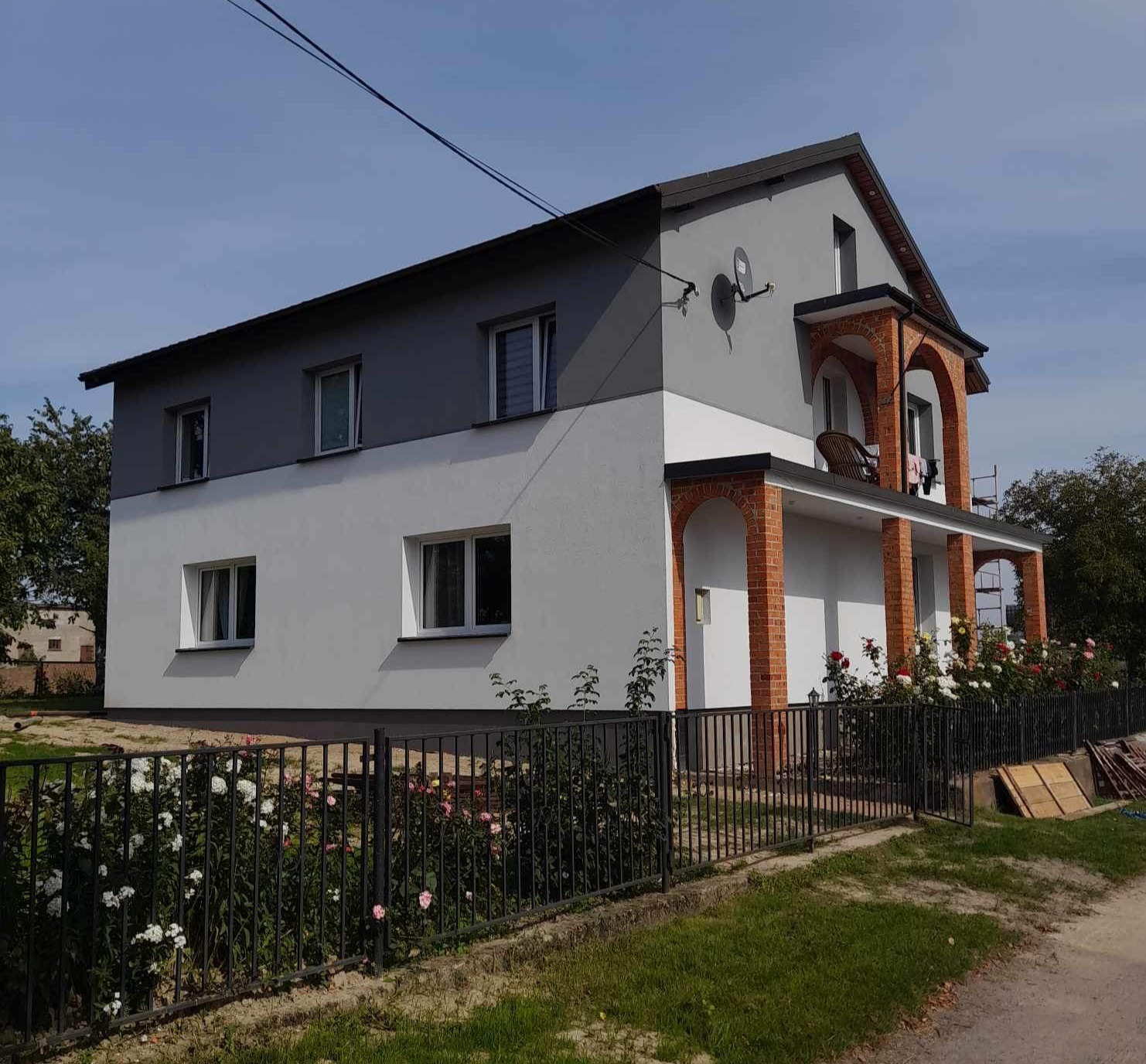 Dwukondygnacyjny dom po remoncie elewacji, z szarą górną częścią i białą dolną, otoczony czarnym ogrodzeniem i kwitnącymi różami. Widoczne ceglane łuki na balkonie.