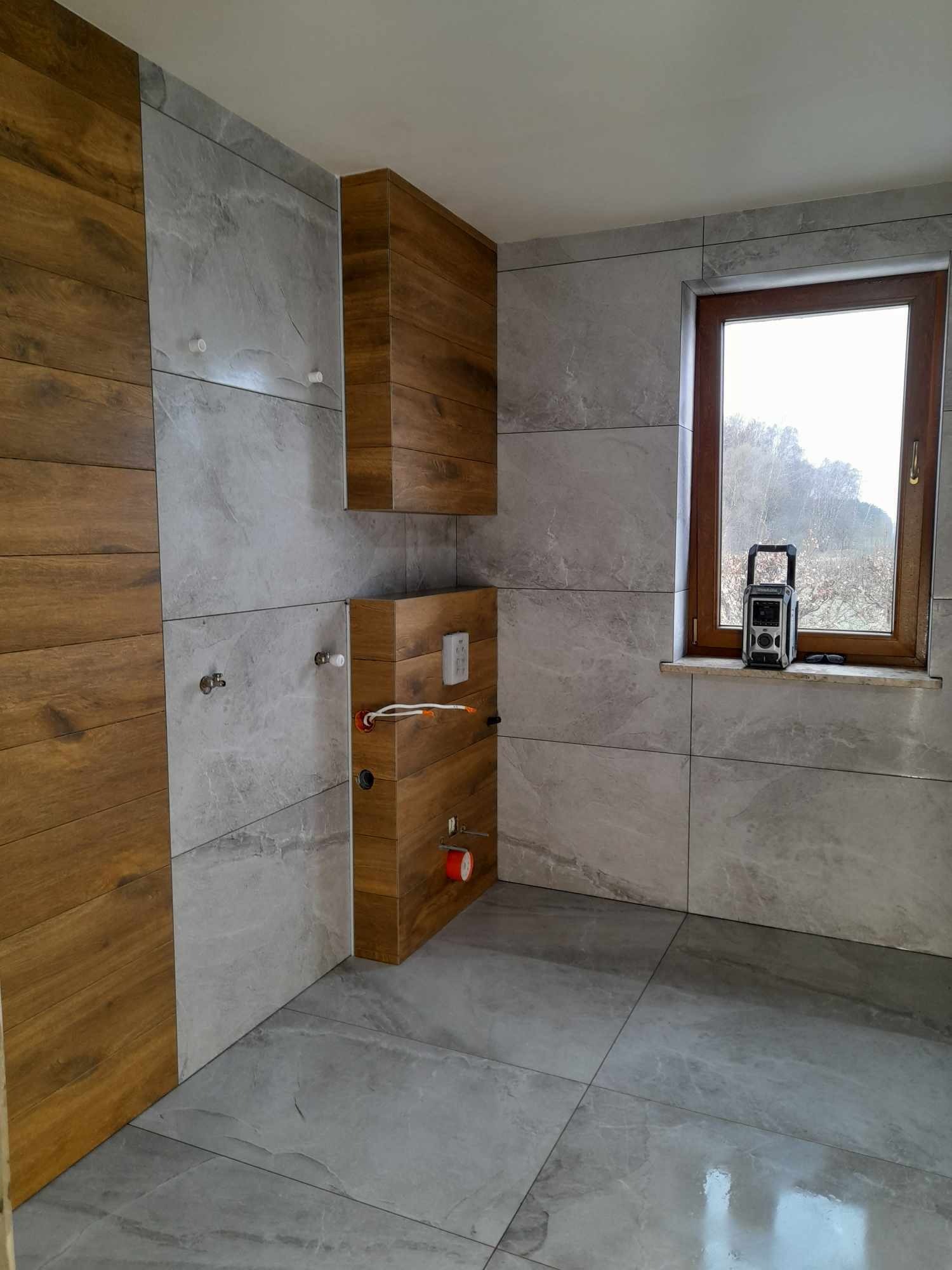 Wykończona łazienka z szarymi płytkami imitującymi marmur i drewnianymi akcentami. Widoczne podłączenia hydrauliczne i okno z radiem na parapecie.