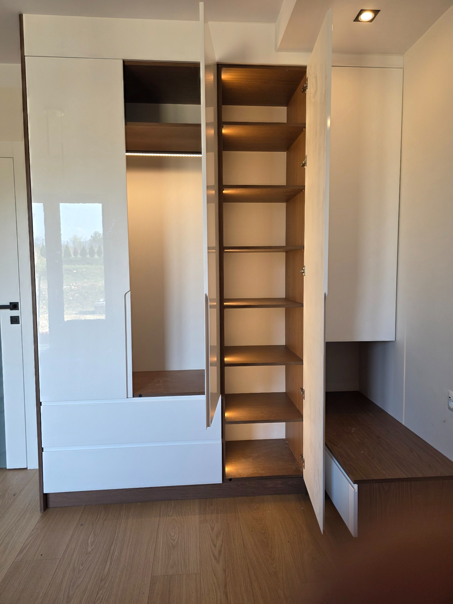 Biała szafa wnękowa z połyskiem i drewnianymi elementami, oświetlona od wewnątrz, z otwartymi drzwiami ukazującymi półki. Widoczny fragment podłogi i ściany.