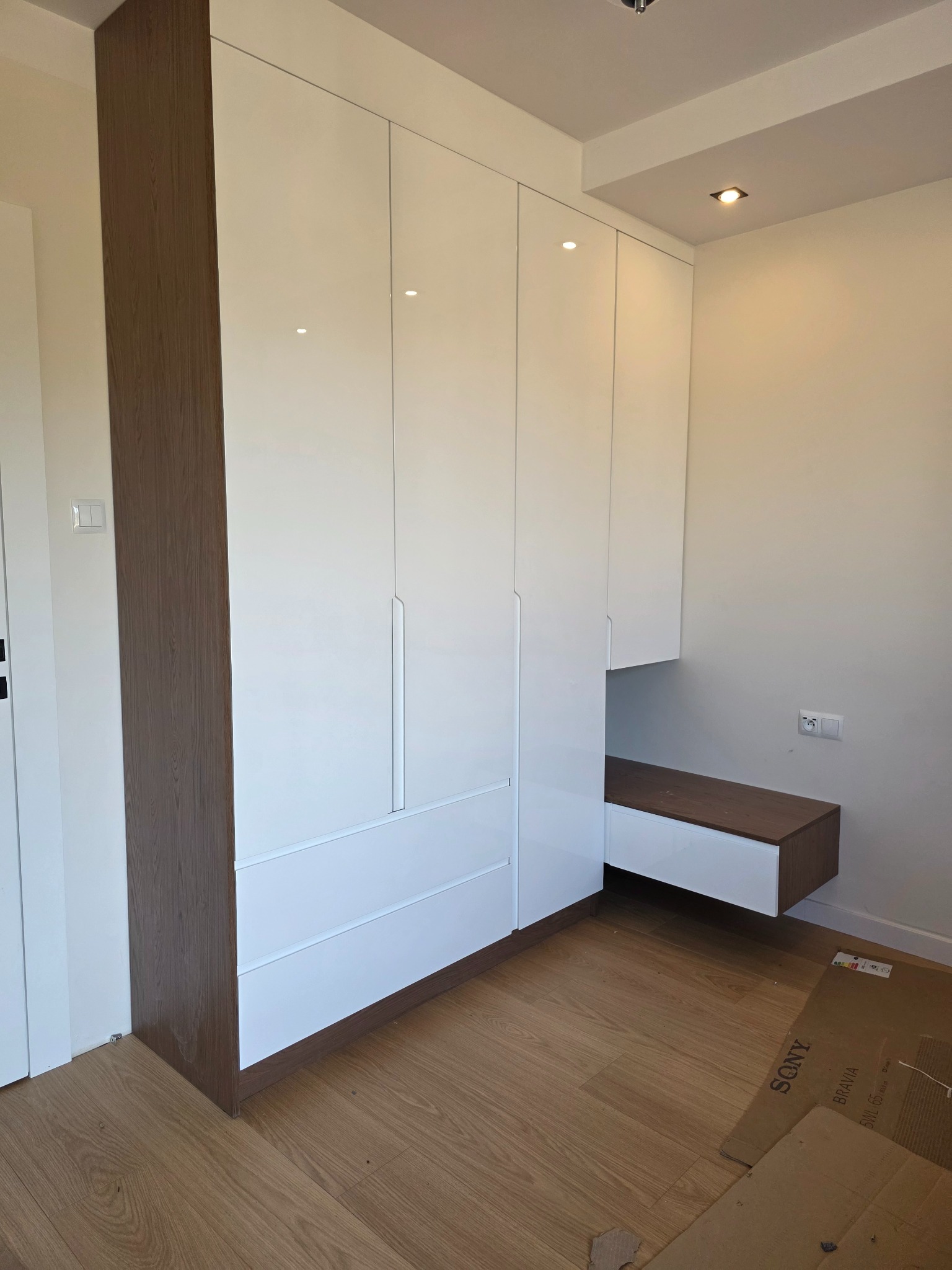 Biała szafa wnękowa z drewnianymi bokami i półką, minimalistyczny design, gładkie fronty, oświetlenie sufitowe, drewniana podłoga, nowoczesne wnętrze.